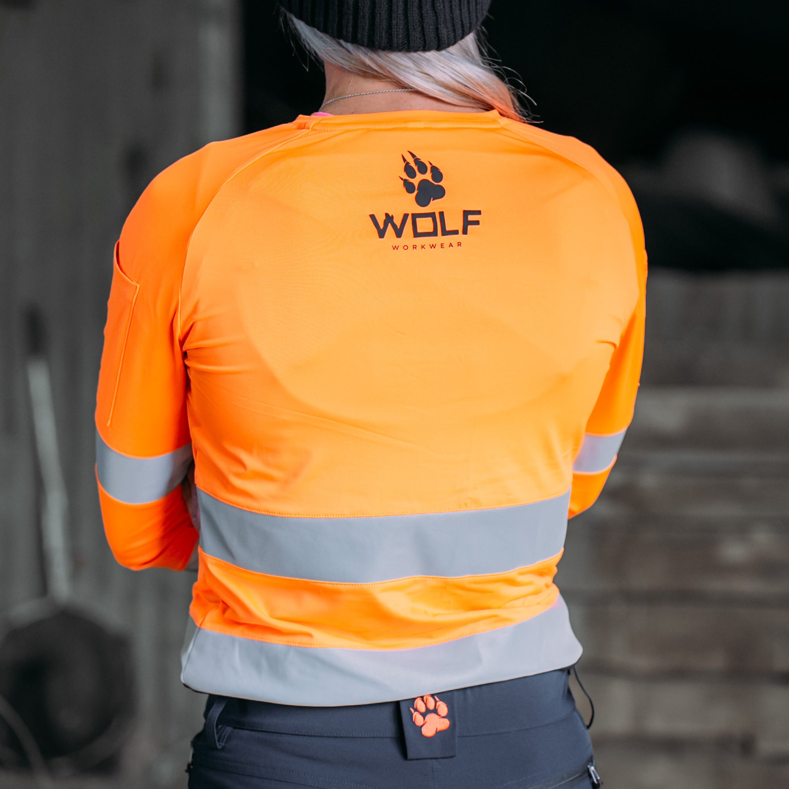 Photo of woman standing in work shed, wearing high visibility orange workshirt.