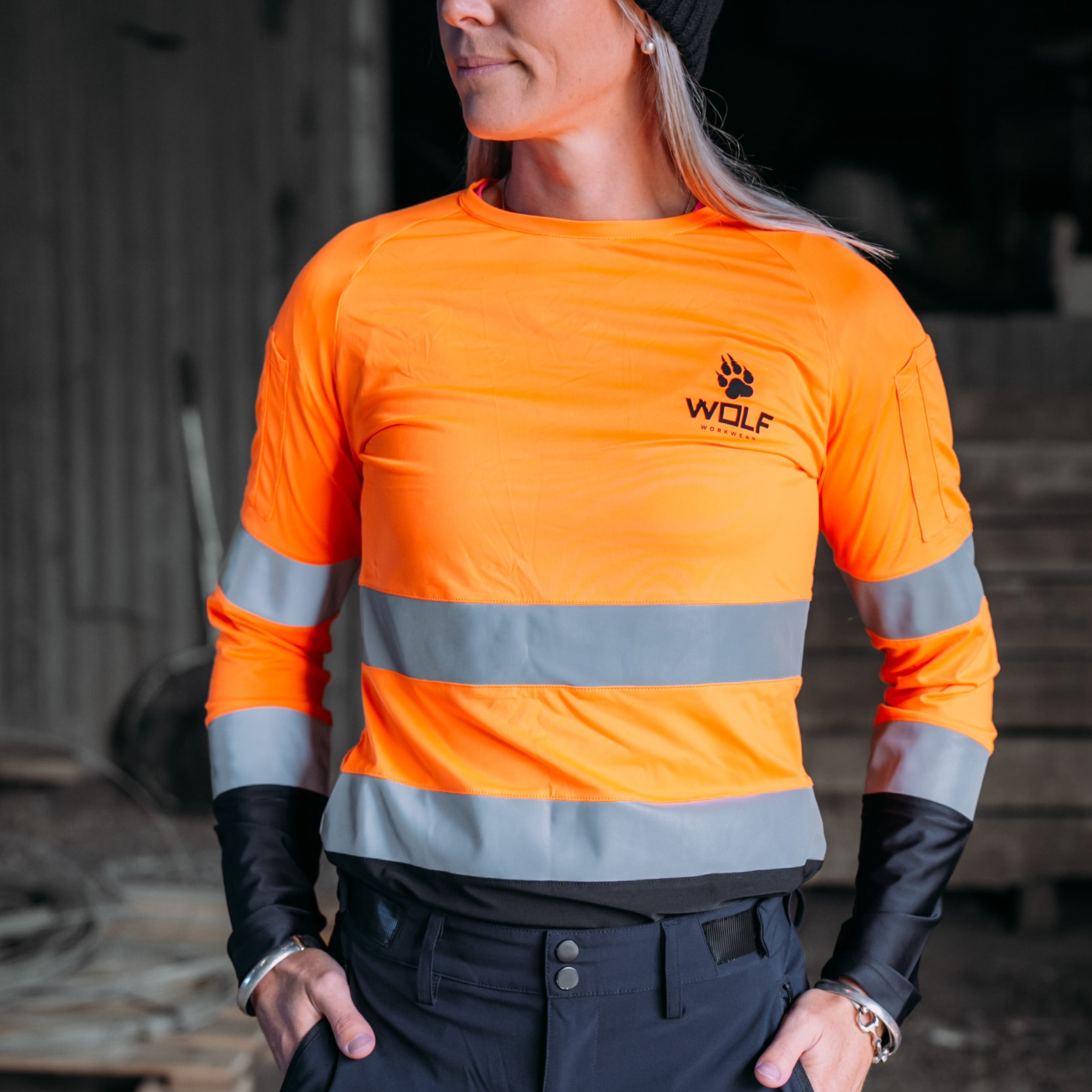 Woman standing in work shed wearing an orange high  visibility work shirt.