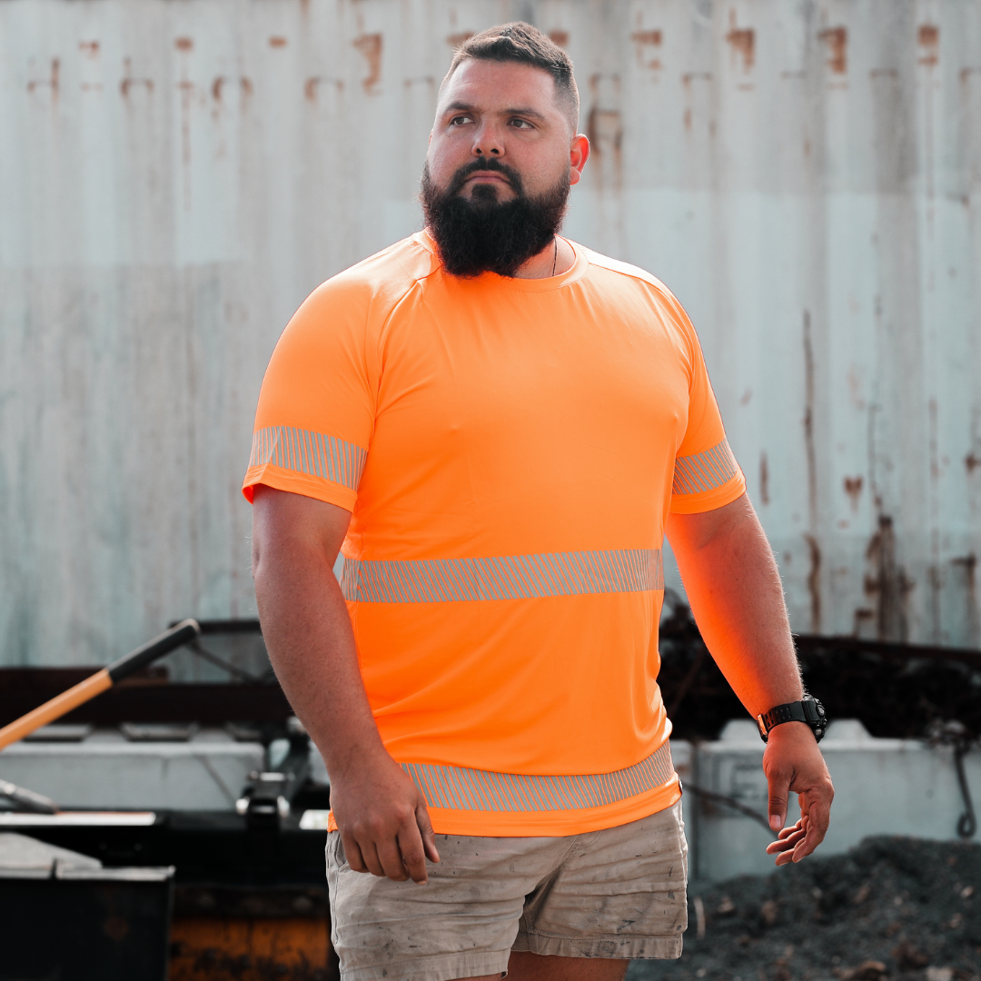 A man wearing an orange high-visibility T-shirt with reflective stripes, standing in front of a blue background with a rugged metal structure.