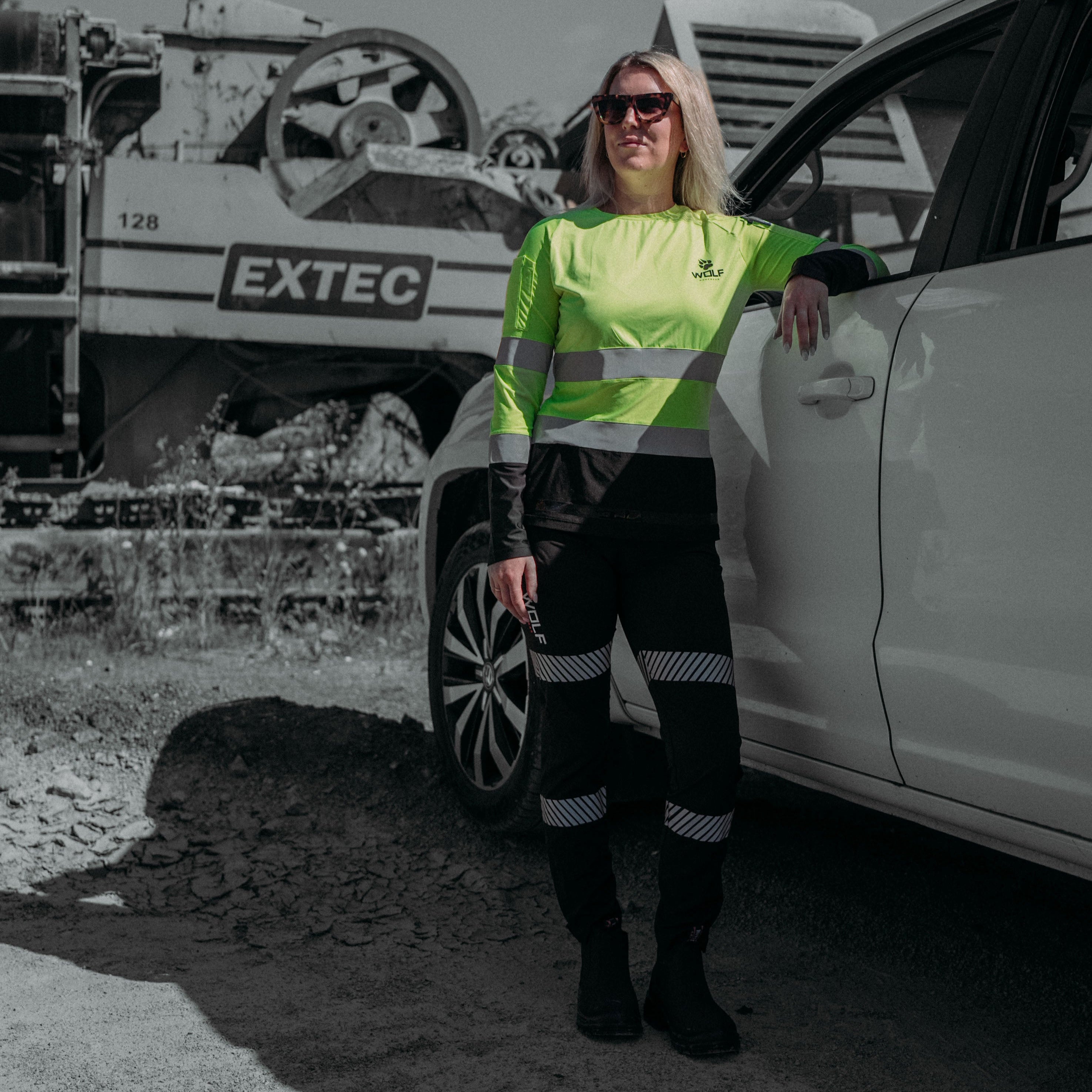 Woman leaning on a car in a worksite, wearing black workpants with high visibility strips.