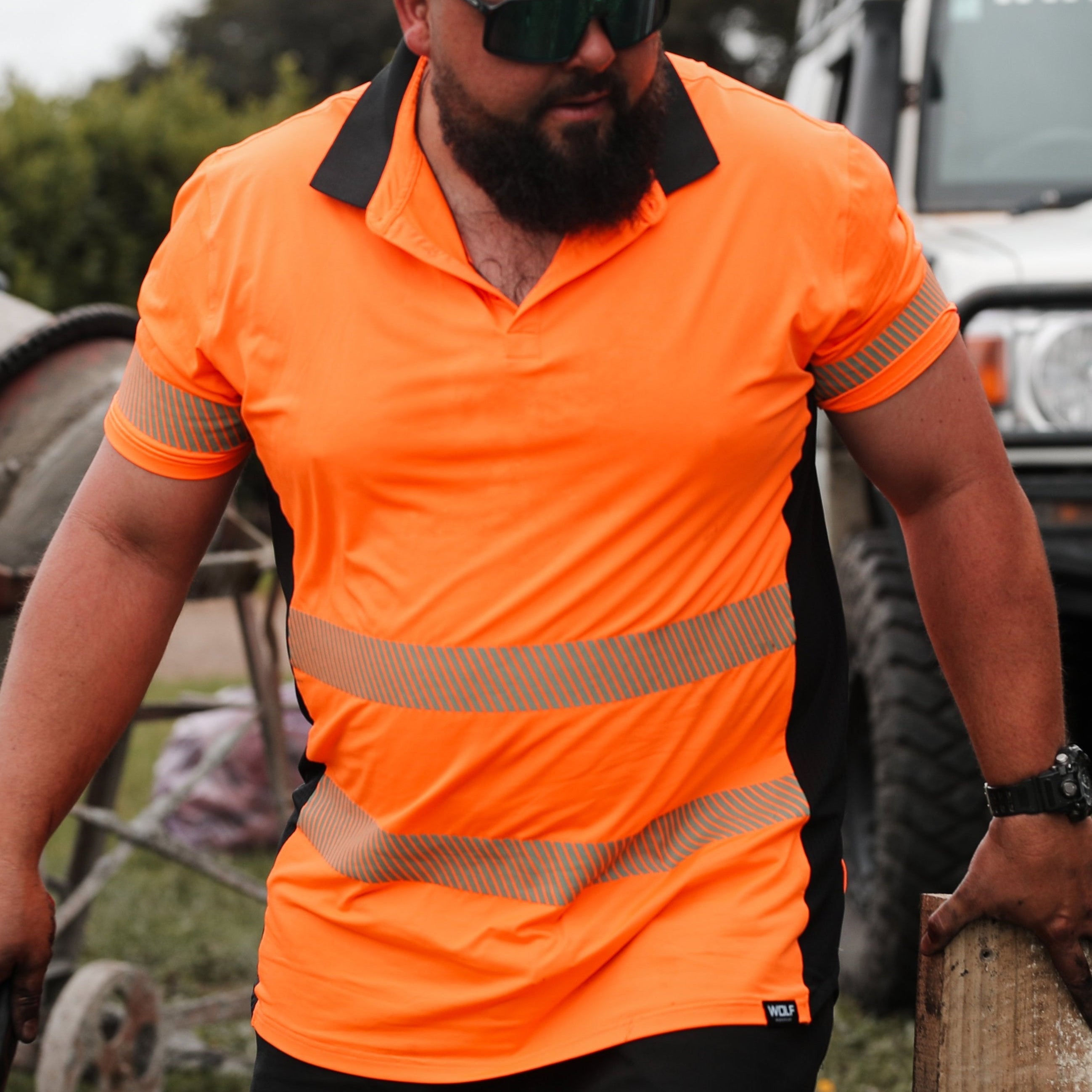 Man walking on work site wearing orange high visibility t-shirt and sun glasses with four wheel drive car and concrete mixer in the background
