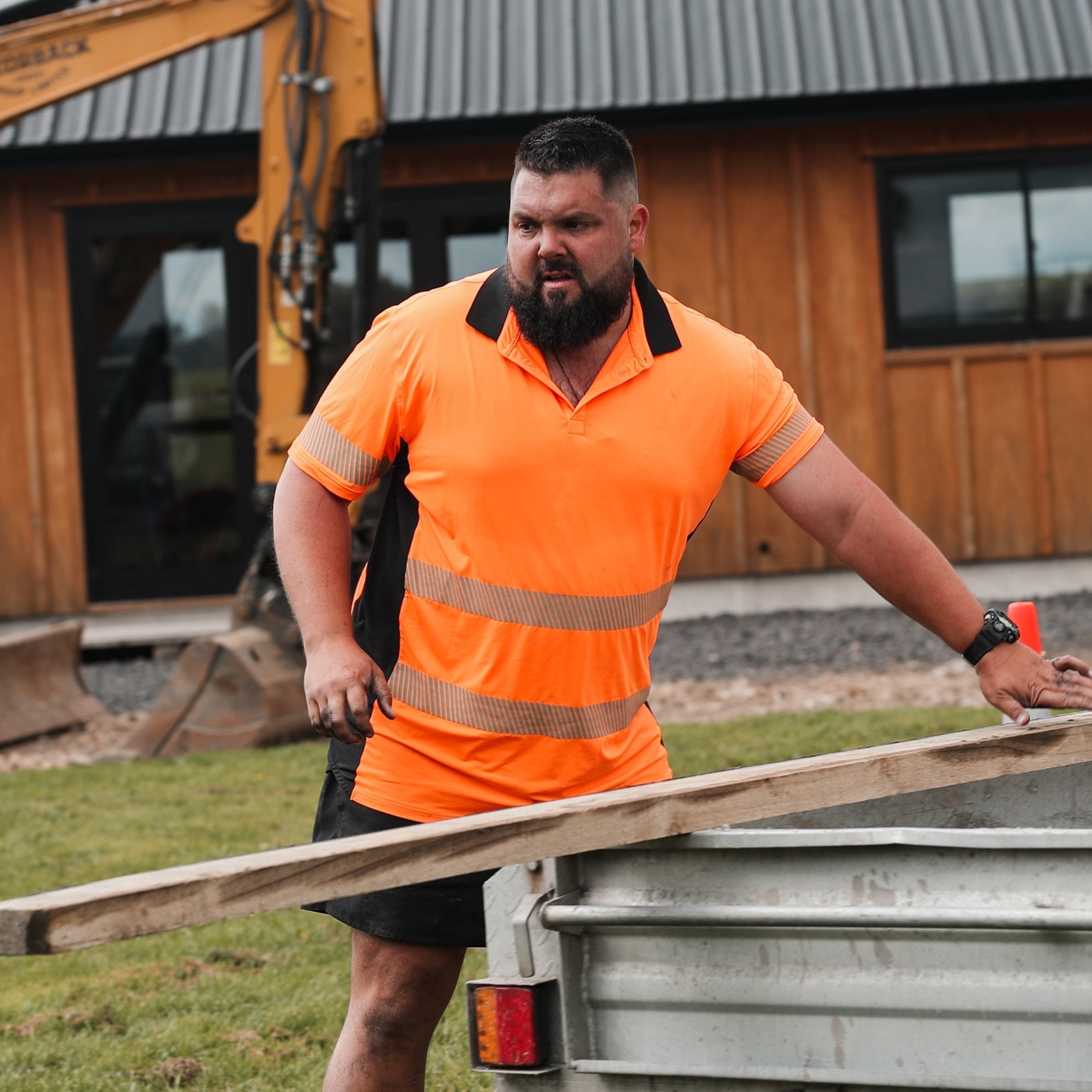 Man on worksite by trailer with digger and house in the background. He is wearing a high visibility work t-shirt