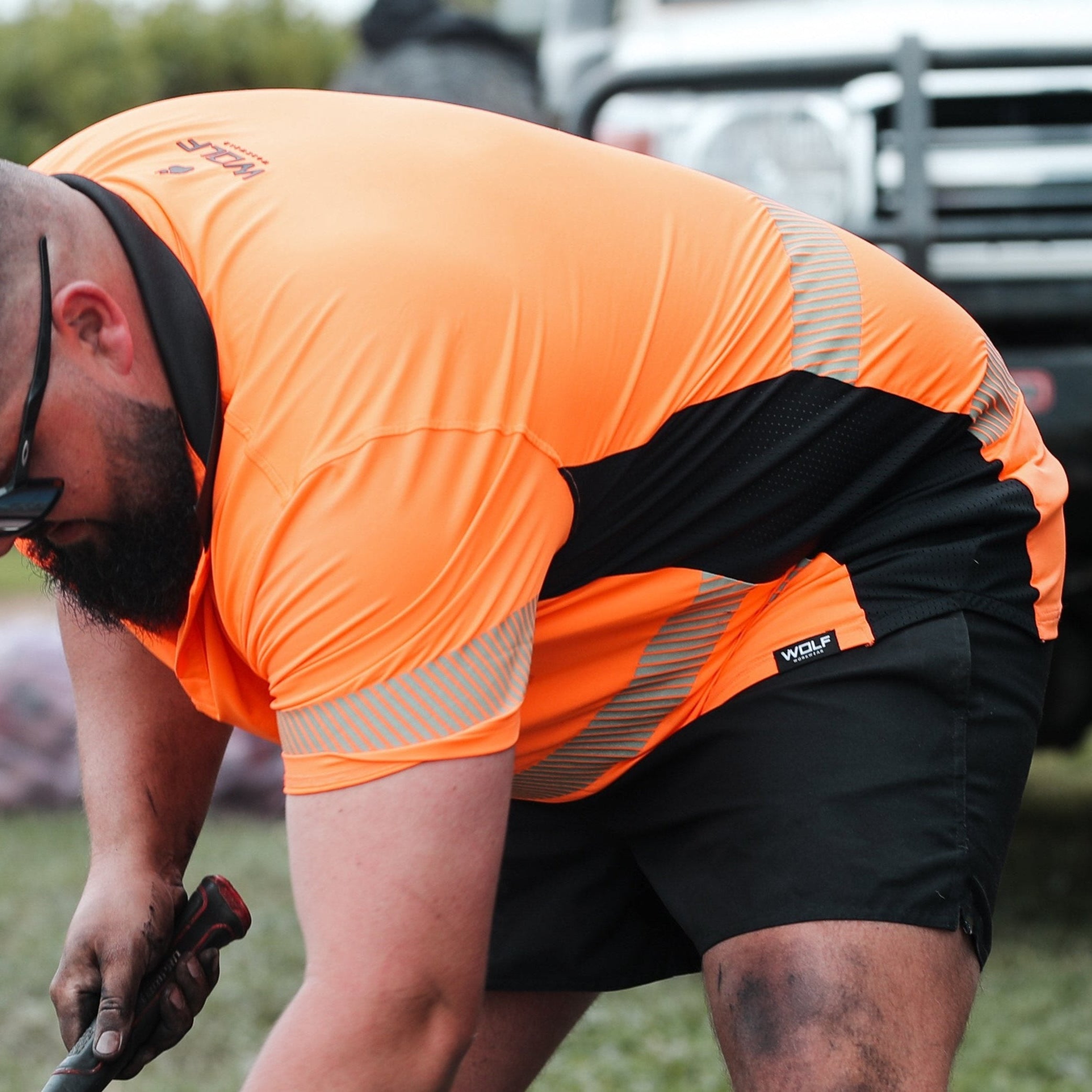 Man on worksite, wearing Wolf Workwear high visibility orange t-shirt, using hammer and wearing sun glassses. There is a concrete mixer and landcruiser in the background.