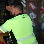 Man in shed, wearing Wolf Workwear High-Visibility Polo Shirt in yellow, climbing into machinery. Up close detail shots of high-visibility strips, Wolf logo on the back and side vents. 