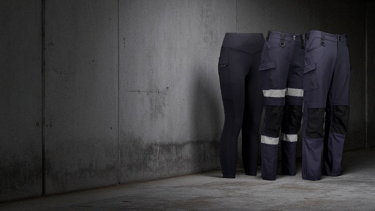 Three pairs of dark blue work pants with reflective stripes against a gray concrete wall.