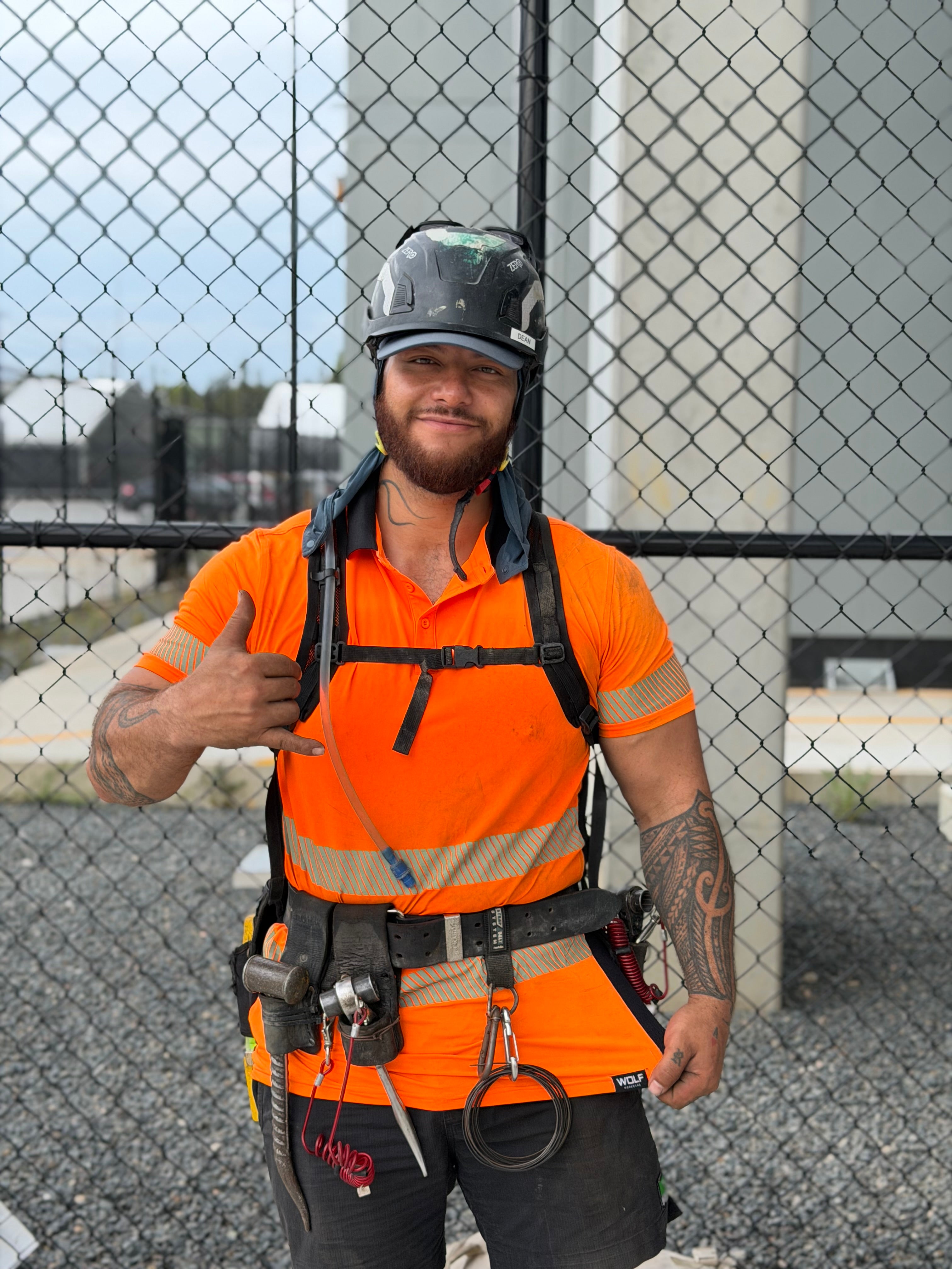 Person in orange safety vest and helmet with climbing gear, giving a thumbs-up in front of a chain-link fence.