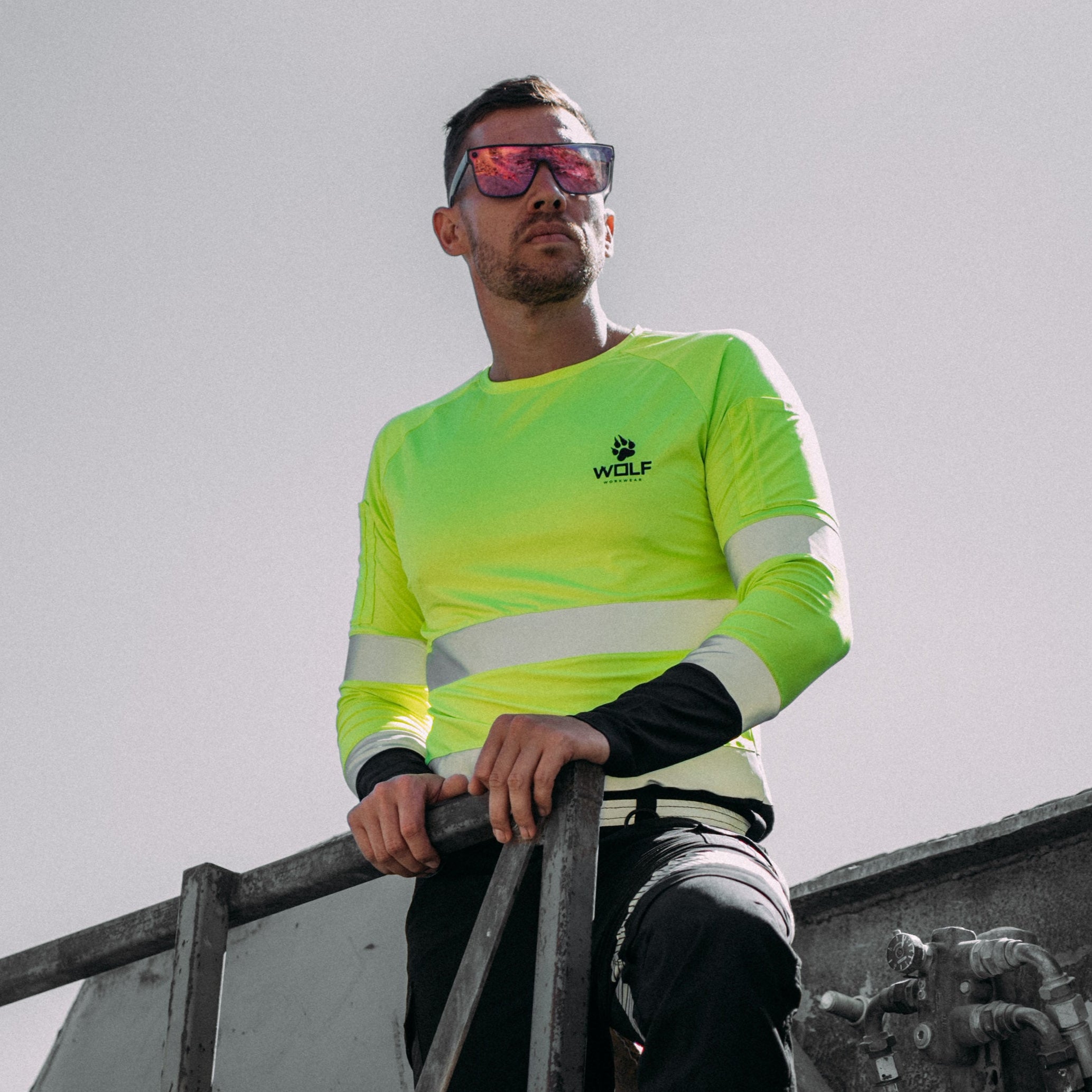 Man standing outside on a work site wearing sunglasses and high visibility long sleeve yellow shirt.