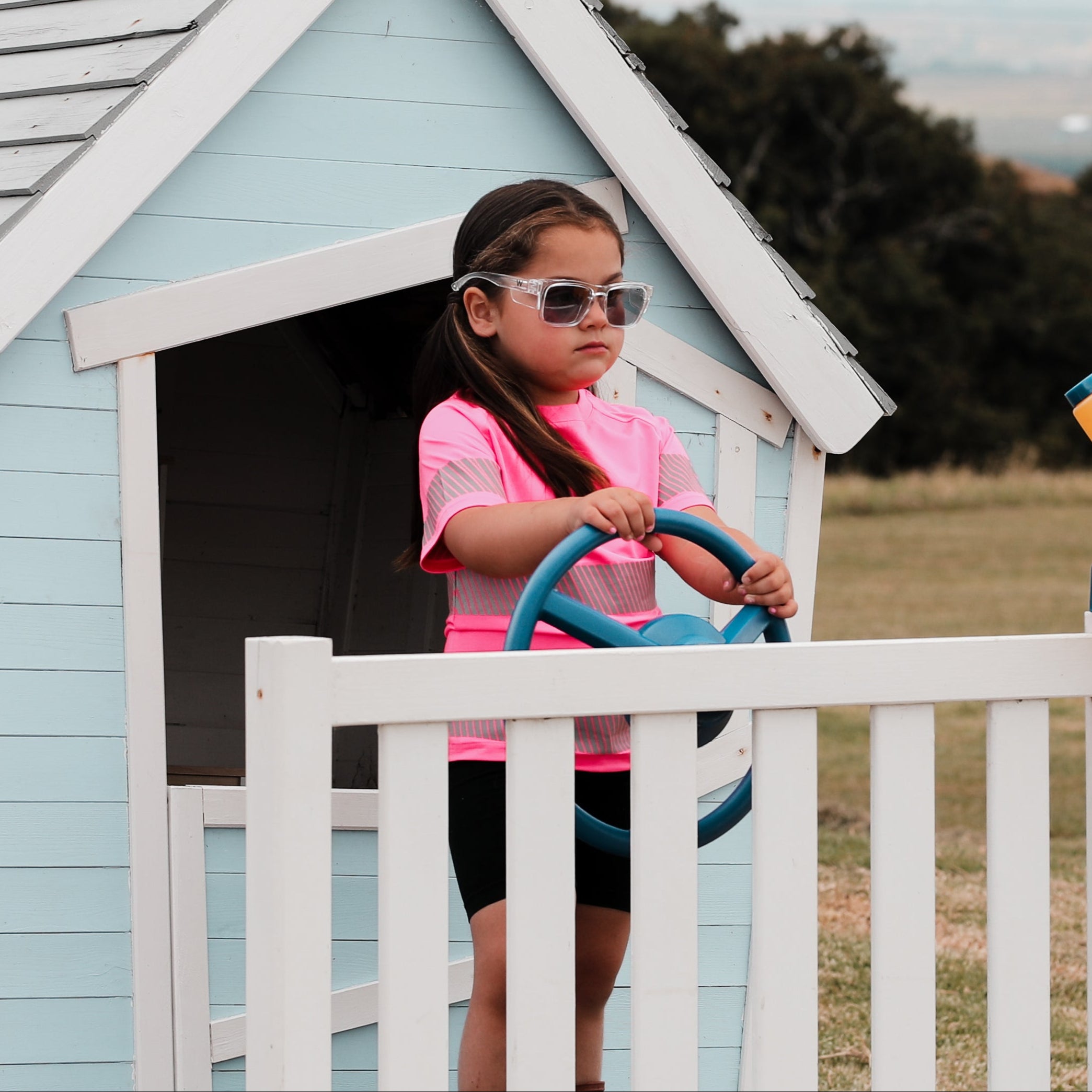 A child standing in a playhouse wearing a pink t-shirt. The playhouse is white with a blue roof.