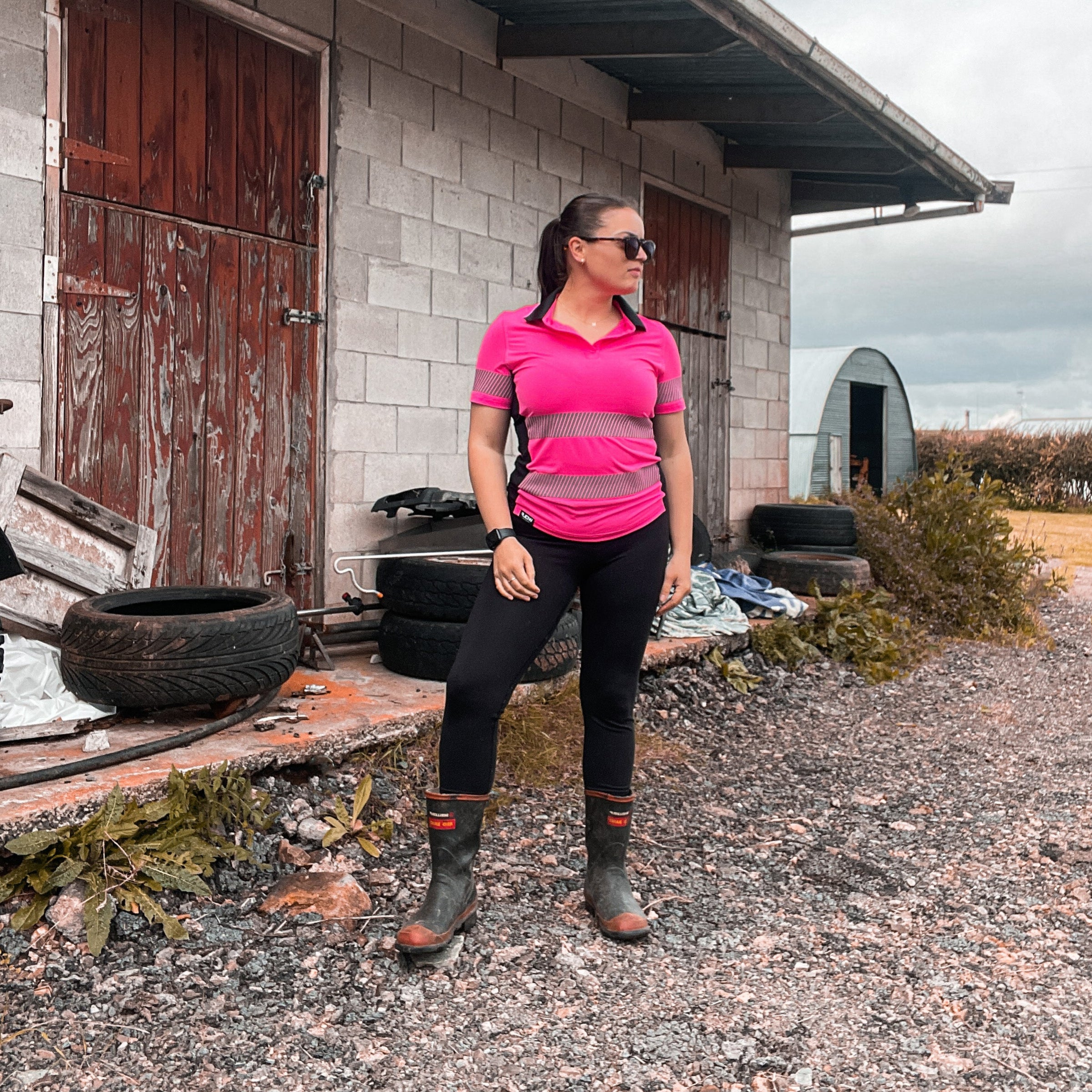 woman standing on rural property wearing pink high visibility polo and gum boots.