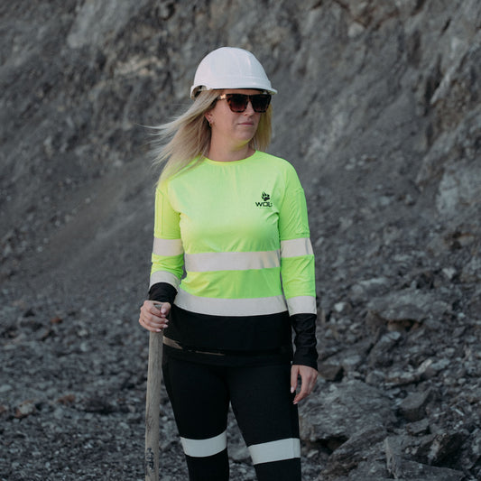 Woman on mining site, wearing hard hat, high visibility long sleeve shirt and work leggings with sun glasses. She is holding a spade.